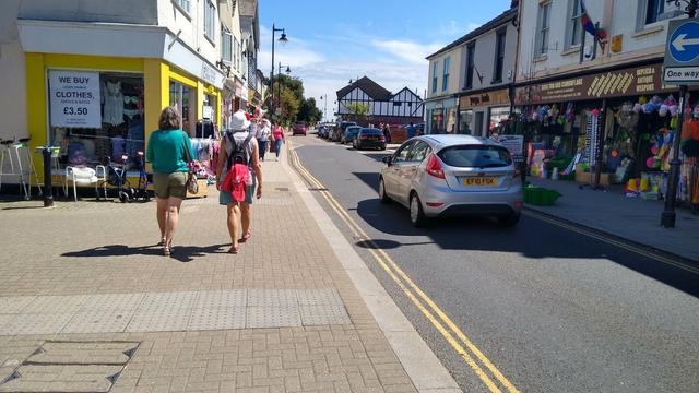 A buff block paved footway continues over a side street with the main road to the right and a footway to the right of that. The street it flanked by shops and the road is one-way heading away from us.

The kerb to the main road carries on over the side road as well, but it is almost flush with the road surface. There is grey blister tactile paving both sides of the junction and two people are walking ahead.