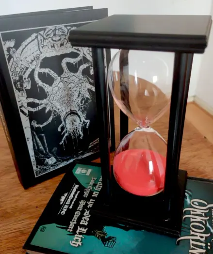 Eine große schwarze Sanduhr steht auf einem Tisch. Der Sand selbst ist rot und soll damit das Fackellicht symbolisieren. Dahinter ist das Shadowdark Regelwerk aufgestellt, auf dem Tisch liegt außerdem das Buch zur Shardowdark Kampagne "Wrath of the Sea Lich".