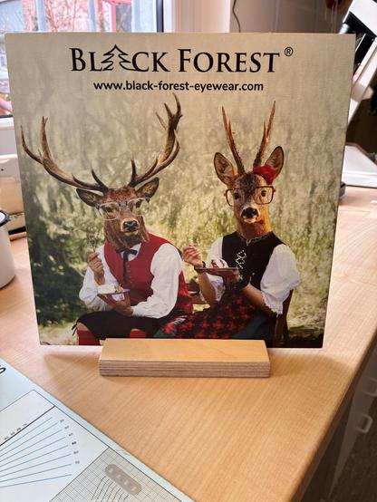 Zwei bebrillte Tier/Mensch-Wesen in schwarzwälder Tracht, die Schwarzwälder Kirschtorte essen. Links ein Männerkörper mit Hirschkopf, rechts ein Frauenkörper mit dem Kopf eines Rehbocks.