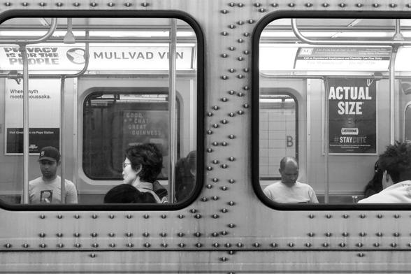 Looking from the platform into the windows of a train stopped at the station. Passengers are visible through the windows, as is the opposite platform. We can see the advertising inside the car as well as the pattern of rivets holding the outside panels together.
