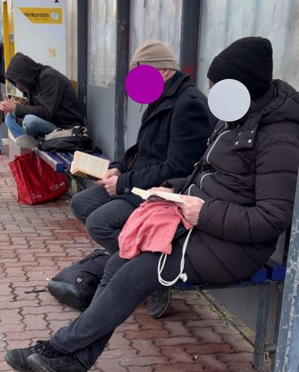 Drei Personen sitzen an einer Haltestelle auf der Bank und lesen hier die ersten zwei Personen haben dunkle Jacken an und schwarze Hosen und eine Mütze. Die dritte Person sitzt etwas weiter weg auf dem zweiten Schneidersitz. Auch sie hat eine dünne Jacke, die Kapuze aufgesetzt und trägt eine Jeans.