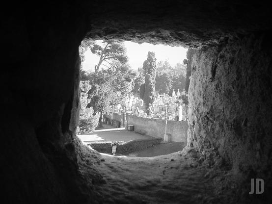 La imagen en blanco y negro muestra la vista a través de una estrecha abertura en una pared de piedra, posiblemente una saetera o ventana defensiva de una fortificación. Al fondo se observa un cementerio rodeado de un muro, con numerosas tumbas y cipreses altos entre los árboles. La escena transmite una sensación de calma y antigüedad.