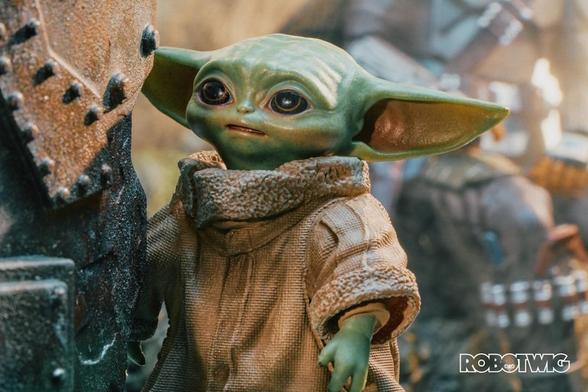 Grogu peers around a bit of old metal. The Mandalorian sits behind him