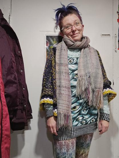 Quinn looking kinda skeptical wearing the woven dataviz scarf, crochet hand spun enby sweater, eyeball sweatshirt, and rainbow pants in a faux knit pattern.