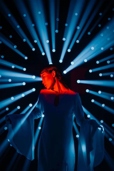 A figure in a flowing white dress poses against a backdrop of blue light beams and circular light spots, creating a dramatic atmosphere.