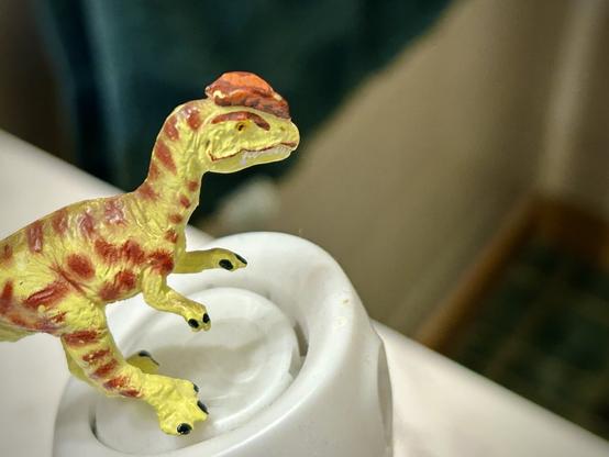 A green Dilophosaurus with brown spots stands on a bottle of air freshener and prepares to erase their shame from the bathroom. A green tile floor is in the far background alongside a white tile wall and a green hand towel hanging off of the rack.