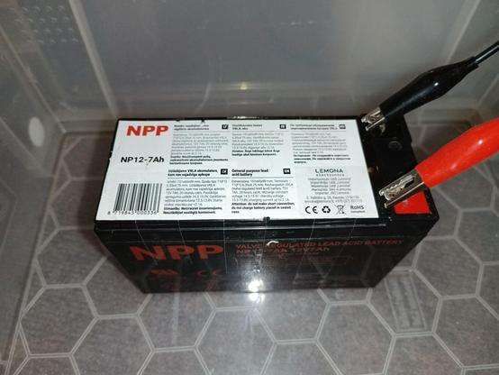 The same battery, with the lid back in place and held by several strips of clear adhesive tape. A red and black crocodile clamp is attached to the poles. It stis in a watertight box.