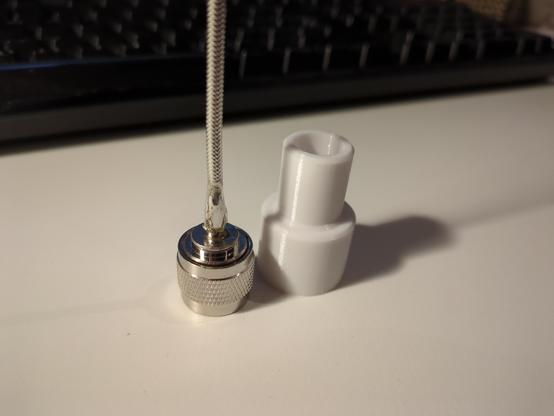 The same silver N-connector soldered to a non-shielded RG402 coax cable but places vertically on a white desk with a black keyboard in background.

Next to it is a white PETG 3D printed adapter, the N-connector will later be glued into to piece to act as a adapter to a PVC conduit which will house the whole antenna.