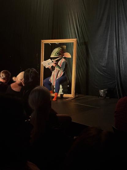 A woman sitting in a picture frame reads a book with a trout mask on her head