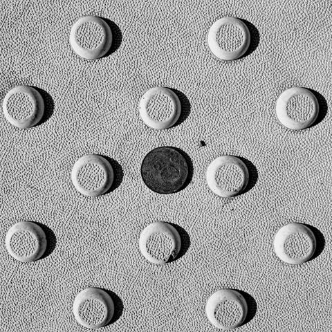 A black and white close-up of a textured surface featuring a grid of large and raised circular bumps, a.k.a. tactile paving. Resting in the center of the grid is a dark gray, oxidized five lira coin. The text on the coin is somewhat dark and obscured due to oxidation and the strong lighting casting distinct shadows from the top left to the bottom right of each raised element. 8a711607-2766-4438-9fb4-8b2c406deaa0