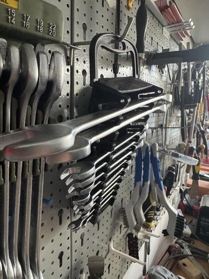 Eine Sammlung verschiedener Werkzeuge wird auf einem Pegboard angezeigt, darunter Schraubenschlüssel, Bürsten und andere Handwerkzeuge. Die Werkzeuge sind ordentlich organisiert und zeigen ihre Metall- und Kunststoffkomponenten vor einem hellen Hintergrund.