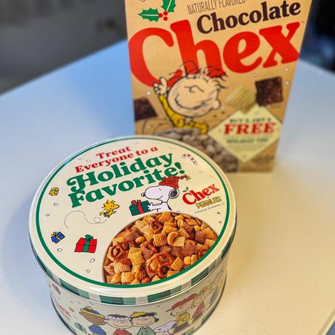 A holiday-themed Peanuts tin filled with Chex mix sits in front of a box of Chocolate Chex cereal. The tin features Snoopy and Woodstock surrounded by presents and candy canes, with text promoting a holiday Chex mix. The cereal box behind it shows a Peanuts character and an offer for a free holiday tin.