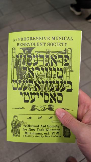 Photo of a hand holding up a neon green paper brochure with text in English and
Yiddish on the cover . It has art of musicians and brass instruments. The text says THE PROGRESSIVE MUSICAL BENEVOLENT SOCIETY, A Mutual Aid Society for New York Klezmer musicians est. 1913
A history zine by Dan Carkner