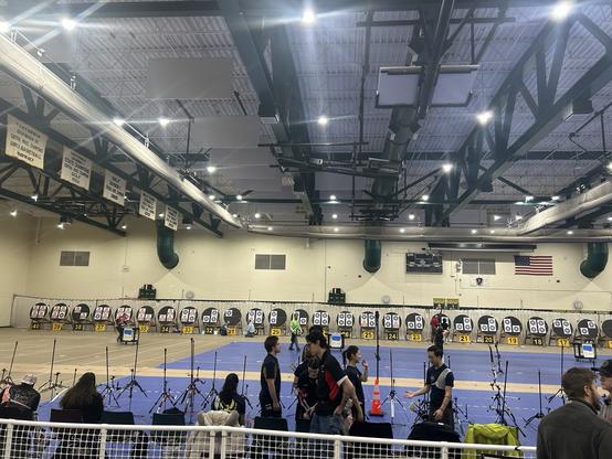 An indoor archery tournament venue photographed from an elevated viewing area. The gymnasium has a high industrial ceiling with exposed beams, ductwork, and bright overhead lighting. Along the back wall, numbered archery target stands (ranging from 17 to 40) are lined up in a row, each displaying black and white bullseye targets. Yellow number markers on the floor indicate shooting positions. The shooting area features blue archery lanes with yellow markings. Championship banners hang on the left wall, and an American flag is displayed on the right wall along with what appears to be a Massachusetts state seal. In the foreground, archers and spectators mill about on the blue floor surface, with archery equipment including bow stands visible. A safety net barrier is visible at the bottom of the frame separating the viewing area from the shooting floor.