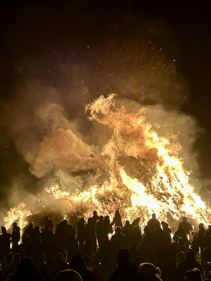 Ein großes Feuer in der Nacht, Biike feiernde Menschen drumherum.