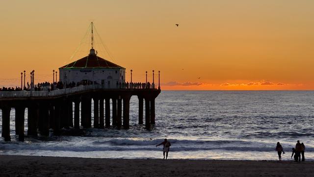 A silhouette of a pier at sunset, with warm orange and yellow hues in the sky. People are gathered on the pier, and a few individuals stroll along the beach. The ocean waves gently lap at the shore.