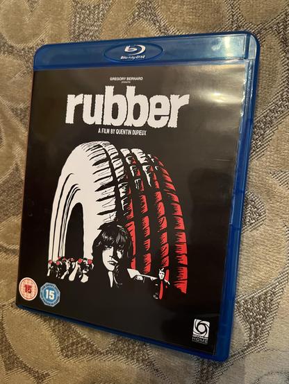 A photo of the case for the blu-ray edition of the movie Rubber by Quentin Dupieux. The cover is mostly black, with a large illustration of a car tyre in white and red. Towards the bottom is an illustration of a black-haired woman, with some people looking into binoculars to the left and a man in a police uniform to the right.