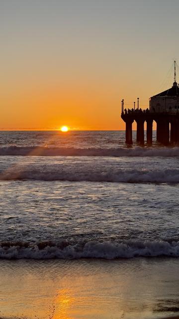 A vibrant sunset over the ocean, with the sun setting just above the horizon. Waves are gently crashing on the shore, and a pier is visible silhouetted against the orange and yellow sky.