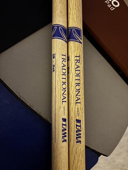 Two wooden drumsticks labeled "Traditional" and "5B - Oak" by the brand Tama, resting on a practice pad with different textures and colors.
