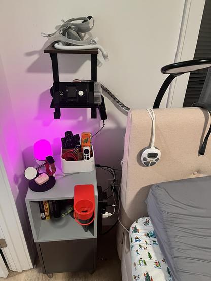 A CPAP machine setup next to a bed, including a mounted mask, tubing, and a machine on a stand, with a bedside table holding electronics, charging devices, and books. Pink glowing lamp on table. Red clamp on cup holder. Black lap behind bed headboard.