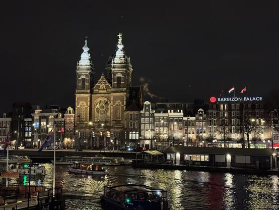 A nighttime view of a historical building featuring twin towers and a large clock, illuminated against a dark sky. In the foreground, boats navigate a waterway reflecting city lights, while nearby buildings include the Barbizon Palace hotel and traditional Dutch architecture.