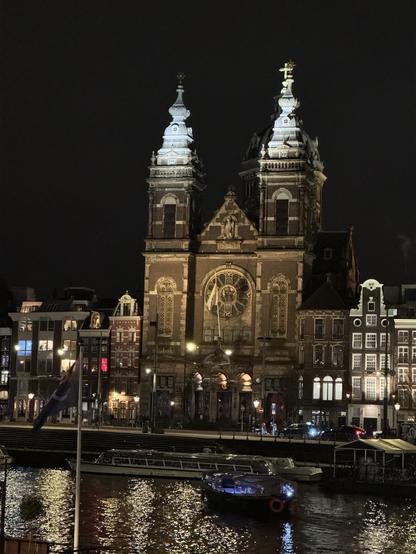 A historic building with two prominent towers illuminated at night, alongside a canal featuring boats. Nearby, traditional Dutch architecture is visible, reflecting in the water.