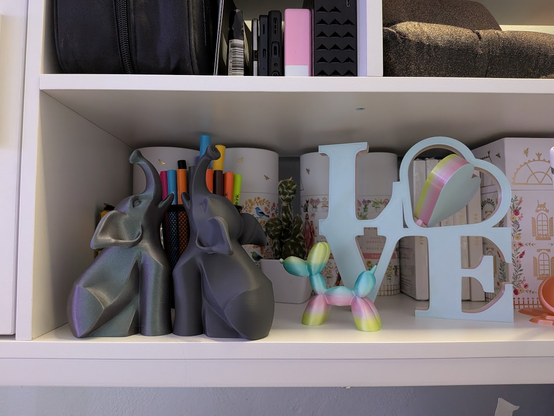 White shelving unit with decorative items including two metallic geometric elephant bookends holding colorful markers, a small pastel balloon dog figurine, a light blue “LOVE” sign with a rainbow heart, floral storage boxes, and a small potted plant in the background.