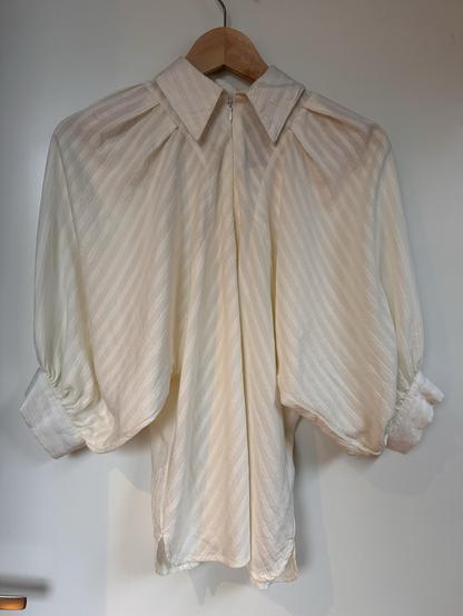An off-white drapy blouse with elbow-length balloon sleeves and a collar. Front closes with a neck-high zipper. Fabric looks like satin and has a subtle stripe pattern that radiates from the neck.