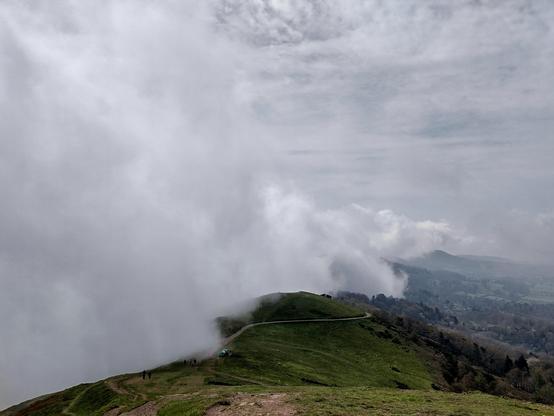 A misty landscape shows a grassy hill with a winding path and a few people in the foreground. Thick clouds envelop the scene, partially obscuring distant hills and trees. The sky is overcast, creating a serene, atmospheric mood.