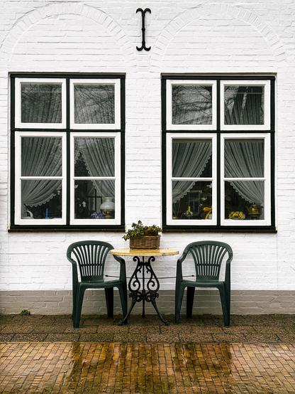A courtyard scene featuring two green plastic chairs and a small round table with a potted plant, set against a white brick wall with two symmetrical windows adorned with sheer curtains. The ground is made of patterned bricks, and the overall ambiance suggests a cozy place.