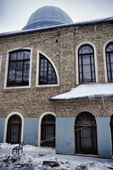 A lonely chair sits stubbornly on the sidewalk in front of an old house. Yellow and red tape is wrapped around it: you're not allowed to walk past. The chair is missing its seat. Icicles hang from the eaves of the house, and there's quite a bit of snow on the street. The house is built of yellowish bricks and is likely over a hundred years old, with large arched windows and a dome on the roof.