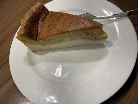 Ein Stück Kuchen auf einem weißen Teller, mit einer goldbraunen Oberkruste und einer leichteren, flauschigen Innenseite. Neben der Platte ruht eine Metallgabel. Der Hintergrund ist eine Holzoberfläche.
