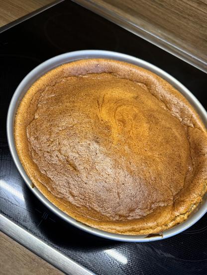 Ein frisch gebackener goldbrauner Kuchen in einer runden Backform, der auf einem schwarzen Herd steht. Der Kuchen hat eine glatte Oberseite und einen leicht erhabenen Rand.