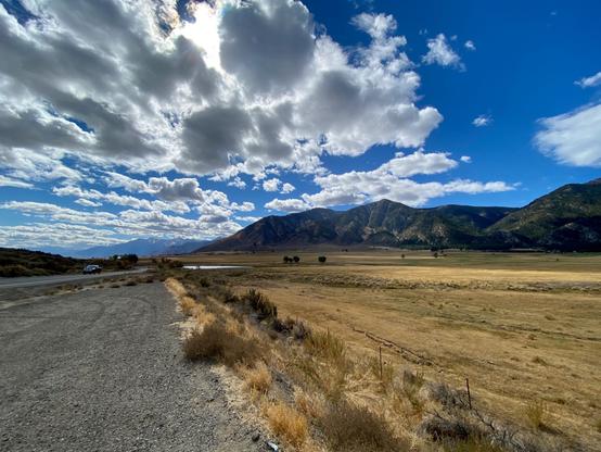 The shot is taken from the side of a road stretching through Jacks Valley/Carson Valley, Nevada. To the right are golden fields with sunlight patches making the gold even brighter. There is a small pond in the distance reflecting the sky, and the entire valley is lined with the Eastern Sierra Mountains which are dark due to the shadows of the amazing clouds above that look like white and gray popcorn scattered against a deep blue sky. The bright gold field against the dark mountains is a dramatic contrast, as are the backlit popcorn clouds. There is more blue sky on the right side of the image.