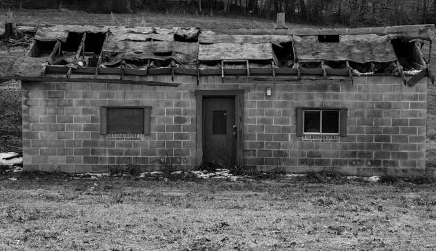 A black and white photo of an small abandoned house of cinder blocks.  Some of the roof is missing and the rest is in poor shape.