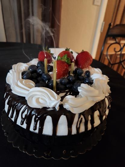 A picture of a white cake with delicious chocolate drizzle and lots of fruit on top, namely blueberries and strawberries. There is a lovely whipped ring around the top of the edge. There are three gold candles, freshly blown out with smoke.