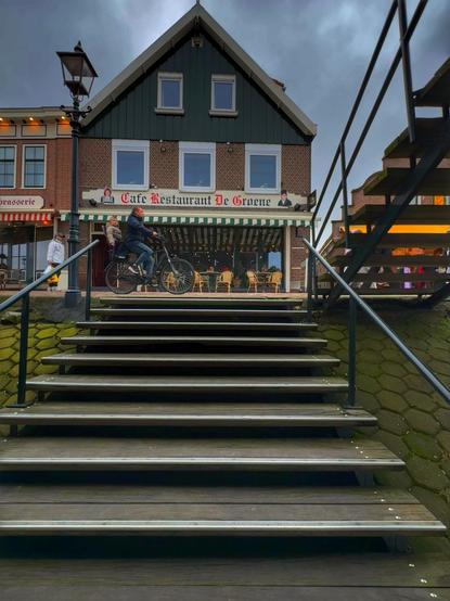 A view of a cafe restaurant titled "De Groene" with outdoor seating, set against a cloudy sky. A person rides a bicycle down a set of wooden steps, leading towards the entrance. The building features a green and brick facade with multiple windows