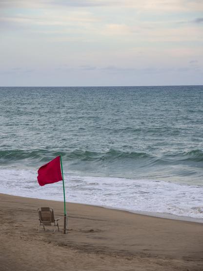 A lifeguard area red flag flies from a pole sticking out from the beach. An empty foldable chair (not the lifeguards’) stands by its side, facing the sea. Surf foam borders the emerald-green water, somewhat choppy from the winds. In the distance the sky is a light grey-blue, with some faint clouds in the distance.