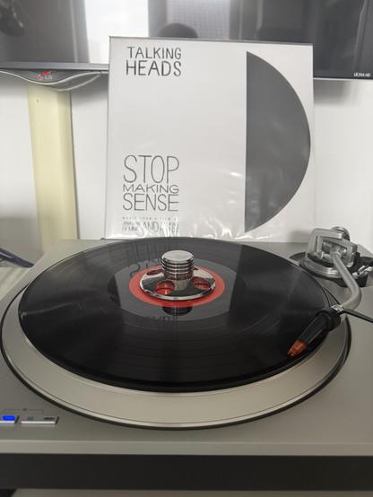 Auf einem silbernen Plattenspieler wird eine schwarze LP abgespielt. Dahinter steht hochkant das Cover. Es ist weiß, im rechten Drittel ist eine stilisierte abgeschnittene Platte. Links steht oben: TALKING HEADS; unten: STOP MAKING SENSE. Darunter ebenfalls in Großbuchstaben: Music from a film by Jonathan Demme and the Talking Heads