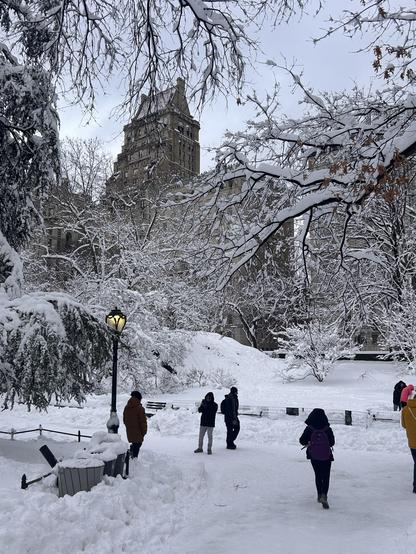 A snowy park scene featuring several people walking along a path. Snow-covered trees and a building are visible in the background, adding to the winter ambiance. Street lamps and benches can also be seen, with snow piled around them.