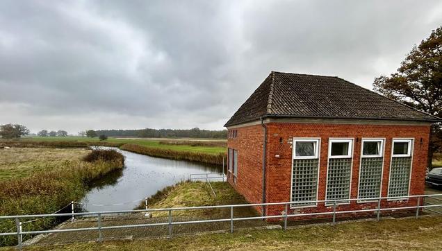 Kleines rotes Gebäude mit Satteldach am Ufer eines schmalen Flusses, umgeben von hohem Gras und einem metallenem Geländer.