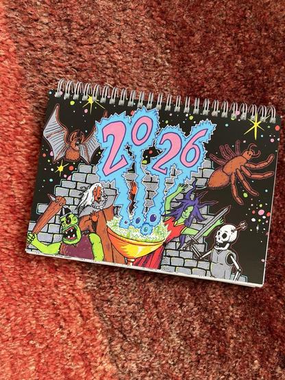 Ein Tischkalender. Das Titelblatt ist mit 2026 beschriftet und wie sehen jede Menge, sehr farbenfrohe Fantasymonster: Skelet, Ork, Riesenspinnne und -Fledermaus. Die Zahl steigt aus einem Zauberkessl hervor, an dem Hexen stehen. Das Ganze wirkt ein bisschen chaotisch, aber nicht zu düstern.