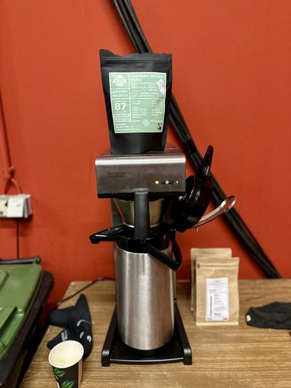 Ein Foto eines Batch-Brewers vor einer roten Wand. Auf der Maschine steht eine Packung Kaffeebohnen. Es ist zu erkennen, dass es sich um Geisha aus Peru handelt