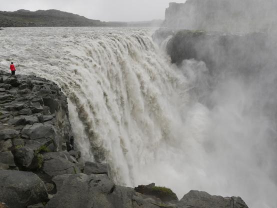 Ein riesiger Wasserfall stürzt tosend zwischen grauen Felsen herab. Oben steht links direkt daneben auf den unwegsamen Felsen eine einzelne Person in leuchtend orangefarbener Jacke und betrachtet den Wasserfall. Von unten steigt eine große Wolke Gischt auf.