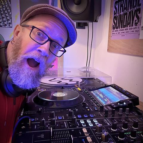 Bearded idiot in a flat cap and glasses staring open mouthed at the camera while leaning over some DJ decks and a mixer