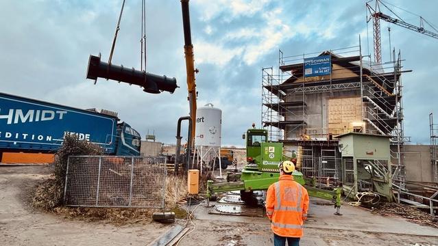 Bauarbeiten: Kran hebt zylindrisches Objekt über Baustelle mit Gerüst und orange gekleidetem Arbeiter. Im Hintergrund Reklame mit Schriftzug "Schmidt".
