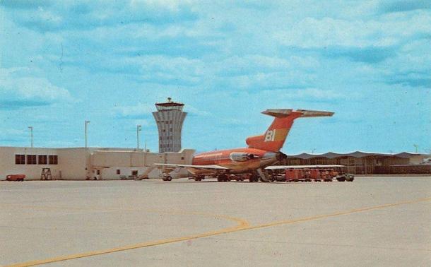 This chrome-era postcard depicts a Braniff International Airways Boeing 727 parked on the ramp at Austin Municipal Airport (Robert Mueller Municipal Airport) in Austin, Texas. The aircraft is shown in Braniff’s distinctive solid-color livery introduced in 1965 as part of the airline’s “End of the Plain Plane” marketing campaign. The vertical stabilizer carries the large white “BI” initials. The aircraft type is a Boeing 727 tri-jet, identifiable by its T-tail configuration and three rear-mounted engines, a model widely used by Braniff for domestic routes during the late 1960s and 1970s.

Ground service equipment, including baggage carts and service vehicles, is positioned near the aircraft’s aft fuselage. The airport’s passenger terminal building appears at left, with jet boarding stairs positioned at the aircraft rather than enclosed jet bridges, reflecting operational practices of the period. Prominently visible in the background is the airport’s flared, hourglass-shaped air traffic control tower, a mid-century modern structure that became a recognizable architectural feature of Austin Municipal Airport prior to its closure in 1999.

Robert Mueller Municipal Airport served as Austin’s primary commercial airport from 1930 until it was replaced by Austin-Bergstrom International Airport in 1999. During the period represented, Braniff International Airways operated extensive service throughout Texas and the United States from this facility.