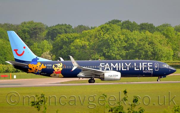 Side view of a twin engined jet airliner taxiing from left to right along a grey runway.
The plane is primarily dark blue, with large white "Family Life" titles on the forward, with much smaller "Hotels" text below the last 2 letters.
A pair of cartoon characters, an orange teddy bear and a yellow dog, are on the rear fuselage, either side of the white registration "G-FDZG".
The tail is a light blue, with a red winky face that looks like the letters "T", "U", and "i" squished together.
Green grass fills the foreground, with trees stretching across the background, under grey sky.