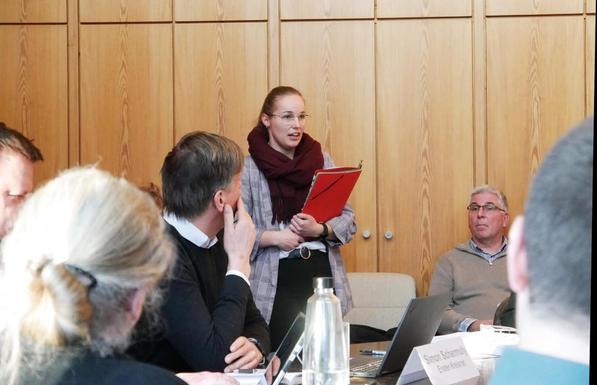 Rote Dokumente haltende Frau spricht vor Publikum in Konferenzraum mit Tischen und Stühlen.