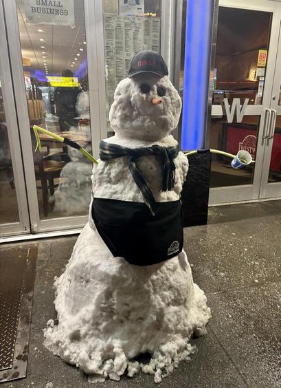 A sidewalk snowman, in front of a NYC diner. He wears a waiter’s apron, has celery arms, and holds a NYC coffee cup for tips.